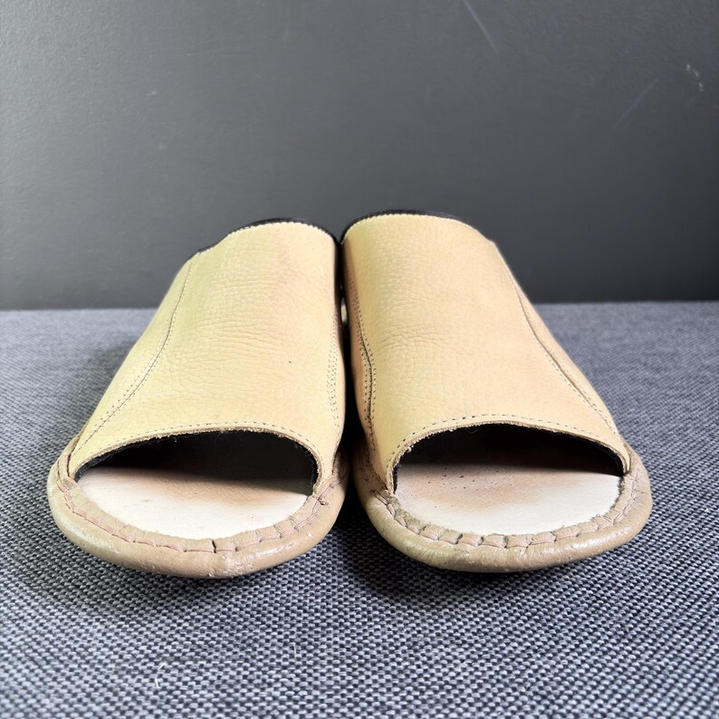 May include: A pair of open-toe slide sandals featuring a light tan, textured leather upper and a smooth, off-white footbed. The edges of the sandals are finished with visible stitching, and the interior lining appears to be a dark, contrasting color.