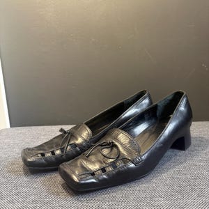 May include: A pair of black leather loafers with a square toe and a small heel. The shoes have a decorative bow and cut-out details on the vamp. The shoes are displayed on a gray surface.