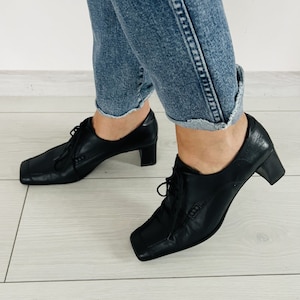May include: Black leather heeled shoes with square toes and lace-up fronts. The shoes have a low block heel.