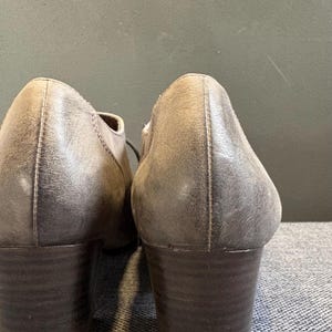 May include: A pair of gray leather heeled shoes. The shoes have a rounded toe and a stacked heel. The leather has a distressed appearance. The shoes are shown from the back.