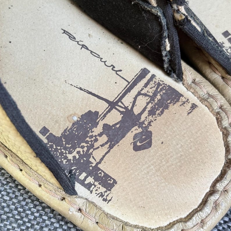May include: A pair of beige sandals with black fabric straps and a black stitched trim. The footbed features a dark brown graphic print of a person surfing and the text Ripcurl in a cursive script, along with a small square logo icon.