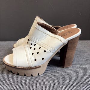 May include: A pair of white leather mules featuring a peep-toe design, a perforated side panel, and a chunky wooden block heel with a thick, beige lugged platform sole. The interior footbed is branded with the text ELENA SHOES MADE IN.