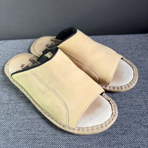 May include: A pair of open-toe slides featuring a textured beige leather upper with black trim along the collar. The footbed is light-colored with visible stitching around the perimeter and a dark graphic logo printed on the heel area of the insole.