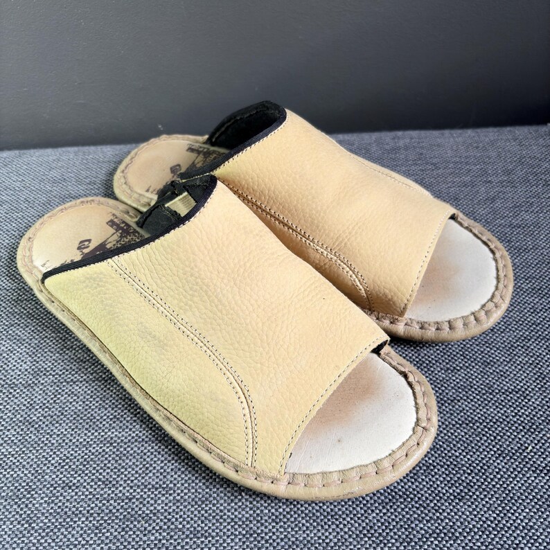 May include: A pair of open-toe slides featuring a textured beige leather upper with black trim along the collar. The footbed is light-colored with visible stitching around the perimeter and a dark graphic logo printed on the heel area of the insole.