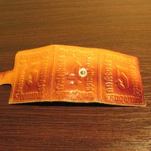 May include: A brown leather wallet with three compartments, each with a decorative design. The wallet has a metal snap closure.