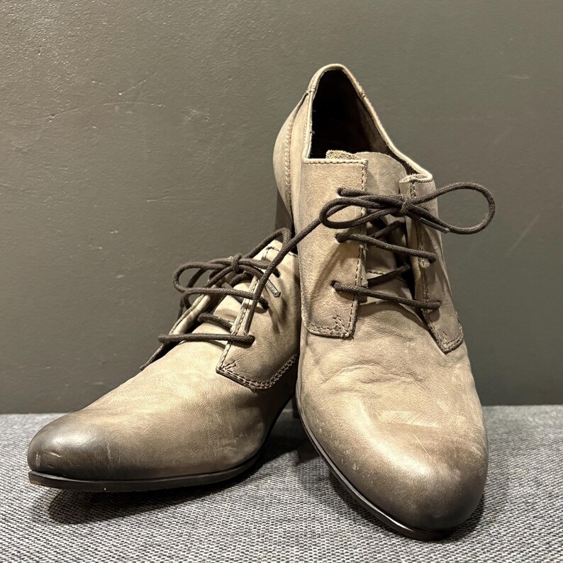 May include: A pair of gray leather lace-up shoes with a stacked heel. The shoes have dark brown laces and a rounded toe. The leather has a distressed appearance, with variations in color.