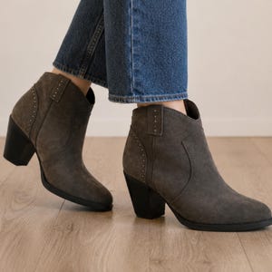 Könnte beinhalten: Graue Wildleder-Ankle Boots mit spitzer Zehenpartie und einem schwarzen Blockabsatz. Die Stiefel haben dekorative Nieten an den Seiten und eine geschwungene Oberlinie. Die Stiefel werden mit einer blauen Jeans getragen.