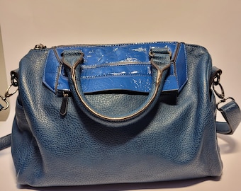 Op de afbeelding: Een blauwe handtas met een getextureerde leren body en een glanzend blauw paneel. De tas heeft zilverkleurige hardware, een handvat aan de bovenkant en een afneembare schouderriem. Het ontwerp omvat een ritssluiting en een voorvak.
