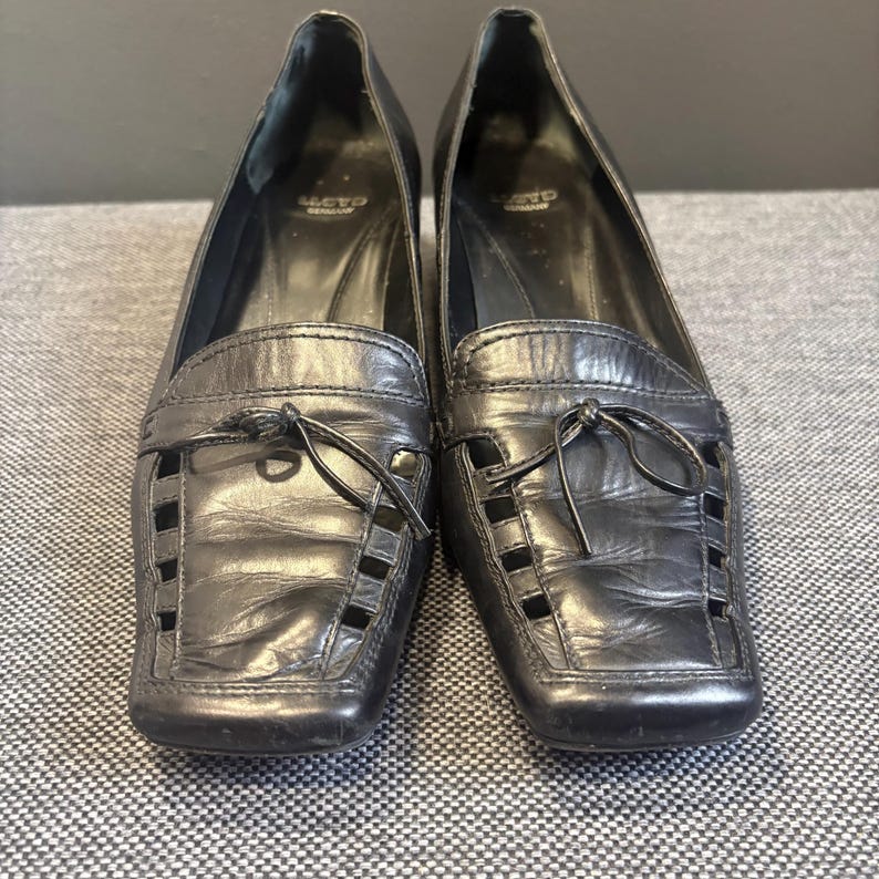 May include: A pair of black leather loafers with a low heel. The shoes have a square toe design with cut-out details and a decorative bow. The brand name "Lloyd Germany" is visible on the inner side of the shoe.
