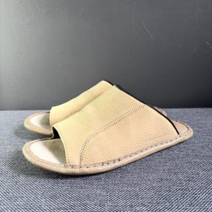 May include: A pair of beige leather slide sandals featuring a textured upper strap with visible stitching, an open-toe design, and a smooth footbed. The edges of the strap are finished with a thin black trim, and the sole is stitched around the perimeter.