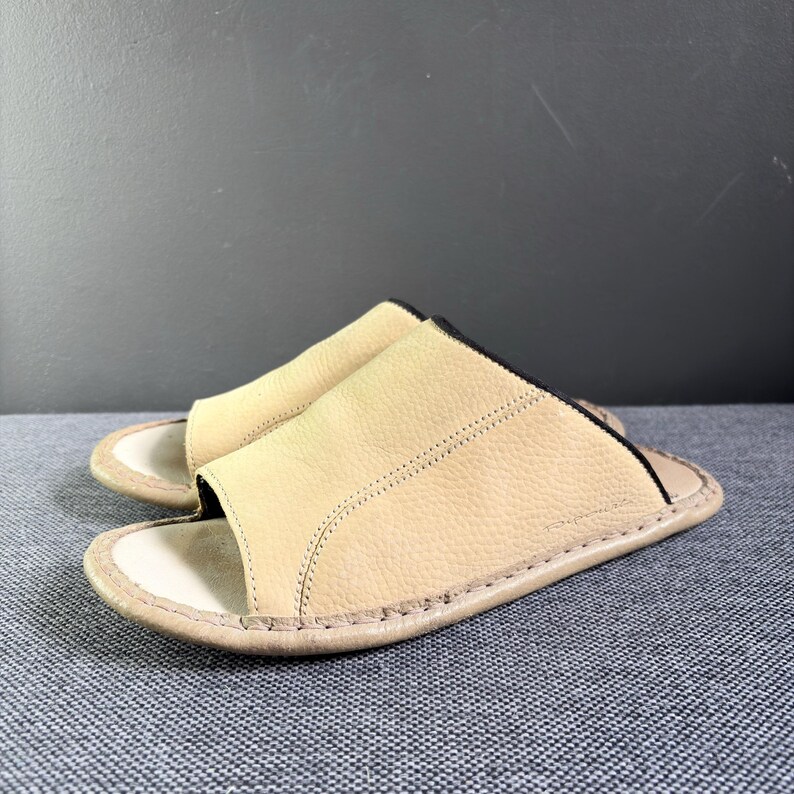 May include: A pair of beige leather slide sandals featuring a textured upper strap with visible stitching, an open-toe design, and a smooth footbed. The edges of the strap are finished with a thin black trim, and the sole is stitched around the perimeter.
