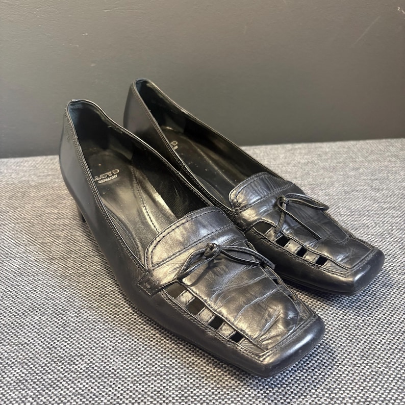 May include: A pair of black leather loafers with square toes and small heels. The shoes have decorative cut-out details on the vamp and small tassels. The brand name "ACTO" is visible on the side of one shoe.