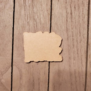 May include: A blank, light brown, wooden cutout in the shape of a state. The cutout has a decorative, uneven edge. The cutout is placed on a wooden surface with vertical wood grain.