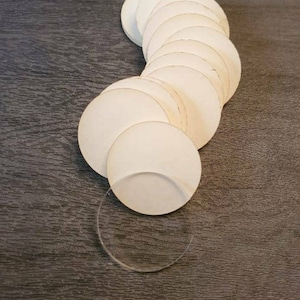 May include: A collection of off-white, circular paper coasters arranged in a curved line. A single, clear, circular coaster is also present. The coasters are on a dark wood-grain surface.