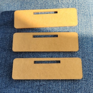 May include: Three rectangular, light brown name tags with a horizontal slot cut out near the top. The tags are made of a smooth material and are arranged on a blue denim surface.