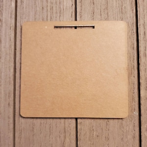 May include: A square, light brown wooden sign with rounded corners and a rectangular slot at the top for hanging. The sign is plain and has a smooth surface.
