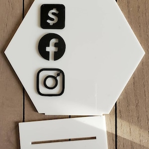May include: A white, hexagonal display stand with black social media icons: a dollar sign, a Facebook logo, and an Instagram logo. A separate white base with a slot is included. The stand is designed to hold a phone or other small items.