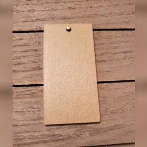 May include: A rectangular, light brown tag with a rounded bottom edge and a small hole at the top. A gold-colored metal eyelet is inserted into the hole. The tag is set against a wooden background.