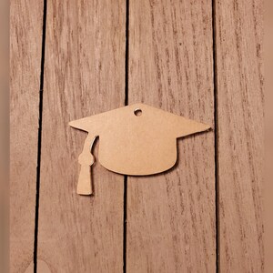 May include: A graduation cap-shaped tag made of light brown cardboard. The tag has a small hole at the top for hanging and a tassel detail. The cap is set against a wooden background.