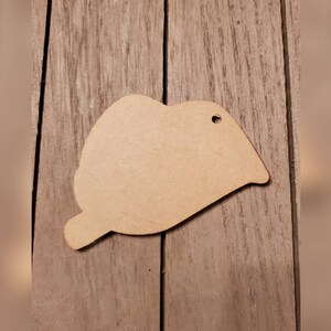May include: A laser-cut, unfinished wooden lip-shaped cutout with a small hole at the top for hanging. The cutout is light brown and rests on a wooden surface. The shape is a stylized representation of a mouth.