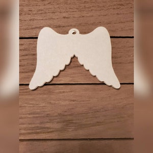May include: A white, wooden angel wing ornament with a scalloped edge. The wings are symmetrical and have a small hole at the top for hanging. The ornament is set against a brown wooden background.