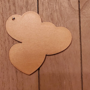 May include: A wooden heart-shaped tag with a cloud-like top. The tag is made of light brown wood and has a small hole at the top for hanging. The tag is lying on a wooden surface.