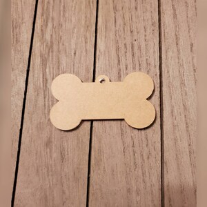 May include: A blank, bone-shaped wooden tag with a small hole at the top for attaching to a pet's collar. The tag is light brown and sits on a wooden surface.