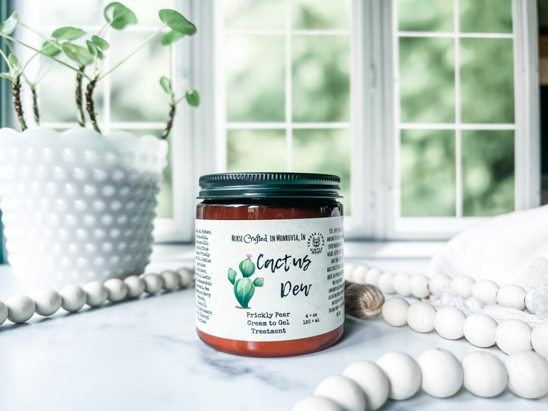 A brown glass jar with a black lid and a white label. The label has a green cactus illustration and the text 'Cactus Dew', 'Prickly Pear Cream to Oil Treatment.