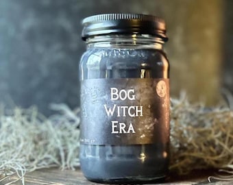 Pode incluir: Um frasco de vidro transparente preenchido com cera cinza-escura, com uma tampa de metal preta. Um rótulo retangular preto na frente apresenta o texto branco "BOG WITCH ERA" em fonte serifada, além de um pequeno logotipo circular.