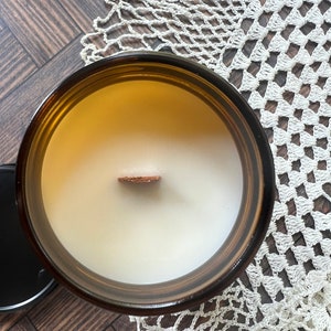 A candle without the lid is photographed from above showing a wooden wick and white wax in an amber glass jar.  Beneath the candle is a chevron wood surface and a white doily.