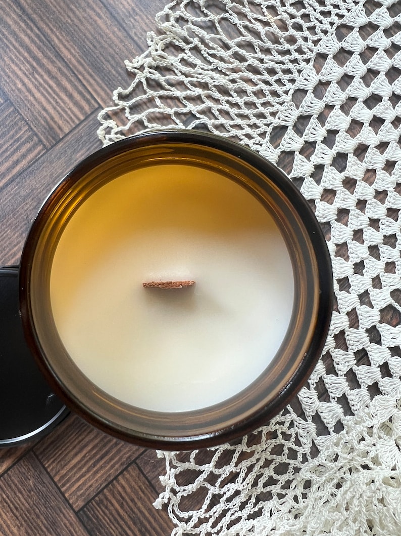 A candle without the lid is photographed from above showing a wooden wick and white wax in an amber glass jar.  Beneath the candle is a chevron wood surface and a white doily.