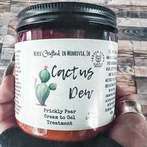 A glass jar with a black lid and a white label. The label says 'NURSE Crafted IN MONROVIA, IN TERESA LYNN'S SIMPLE Cactus Dew Prickly Pear Cream to Gel Treatment and has a green cactus illustration.