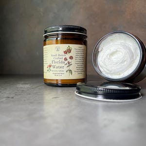 Pode incluir: Um frasco de vidro castanho de manteiga corporal batida com tampa preta e r&oacute;tulo cor de creme com a inscri&ccedil;&atilde;o Small Batch Crafted Florida Water. O r&oacute;tulo apresenta ilustra&ccedil;&otilde;es florais vermelhas. Um segundo frasco aberto revela creme branco.