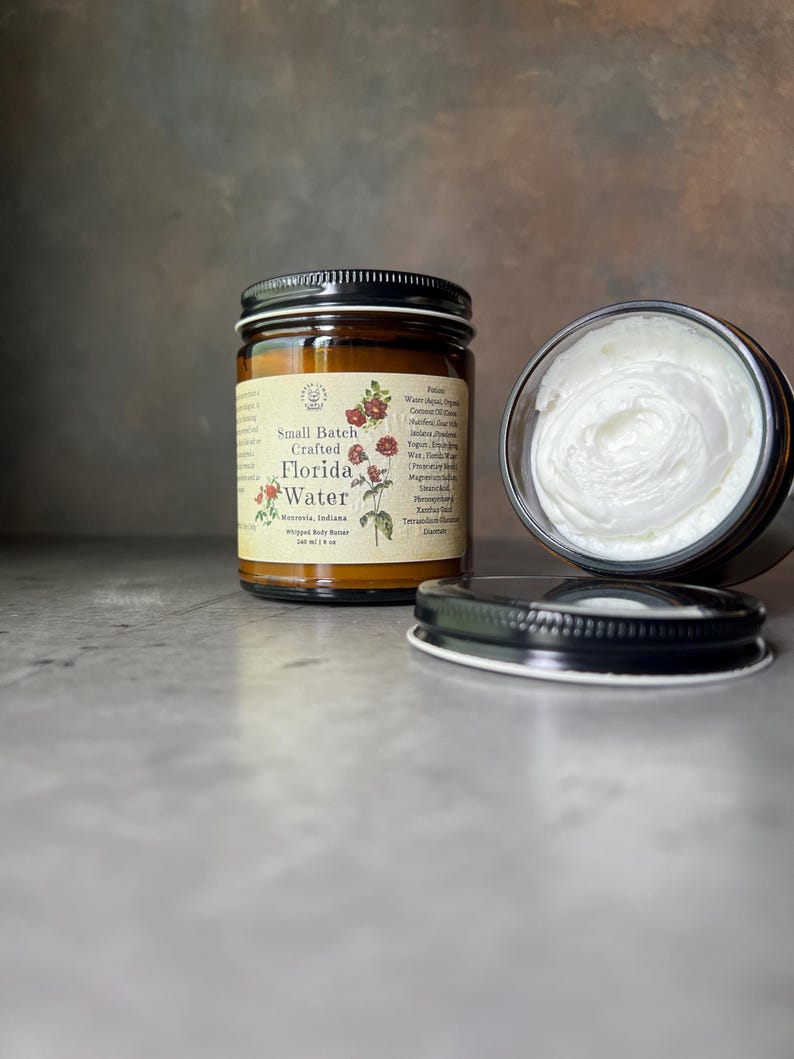 Pode incluir: Um frasco de vidro castanho de manteiga corporal batida com tampa preta e r&oacute;tulo cor de creme com a inscri&ccedil;&atilde;o Small Batch Crafted Florida Water. O r&oacute;tulo apresenta ilustra&ccedil;&otilde;es florais vermelhas. Um segundo frasco aberto revela creme branco.