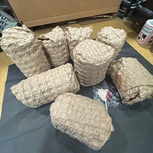 Pode incluir: Sete objetos cil&iacute;ndricos embrulhados em papel castanho com textura de favo de mel. Os itens est&atilde;o dispostos em grupo sobre uma superf&iacute;cie escura. Um pequeno chupa-chupa em forma de cora&ccedil;&atilde;o em pl&aacute;stico transparente est&aacute; entre os itens.