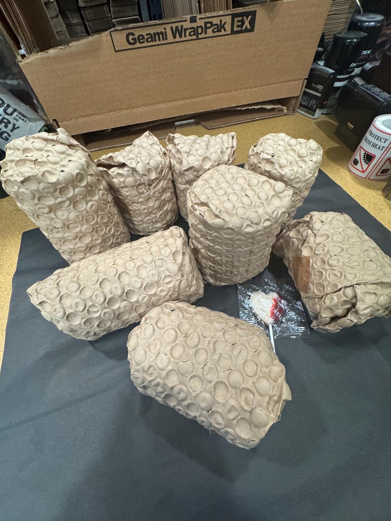 Pode incluir: Sete objetos cil&iacute;ndricos embrulhados em papel castanho com textura de favo de mel. Os itens est&atilde;o dispostos em grupo sobre uma superf&iacute;cie escura. Um pequeno chupa-chupa em forma de cora&ccedil;&atilde;o em pl&aacute;stico transparente est&aacute; entre os itens.