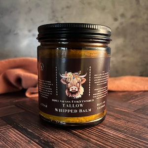 May include: A glass jar with a black lid filled with a yellow balm. The label reads "100% Grass Fed/Finished Tallow Whipped Balm" and has a cute illustration of a Highland cow with a floral crown. The best by date is 12/2024.