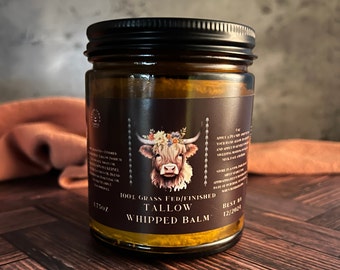 May include: A glass jar with a black lid filled with a yellow balm. The label reads "100% Grass Fed/Finished Tallow Whipped Balm" and has a cute illustration of a Highland cow with a floral crown. The best by date is 12/2024.