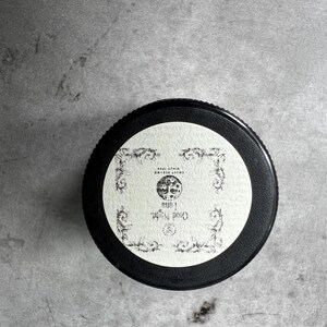 Op de afbeelding: Een ronde zwarte deksel van een potje met een cr&egrave;mekleurig etiket met sierlijke zwarte filigraanranden. Het etiket toont een centrale illustratie van een maan, de tekst "Good Night Luna" en de kleine tekst "SWEET DREAMS SINCE 1998".