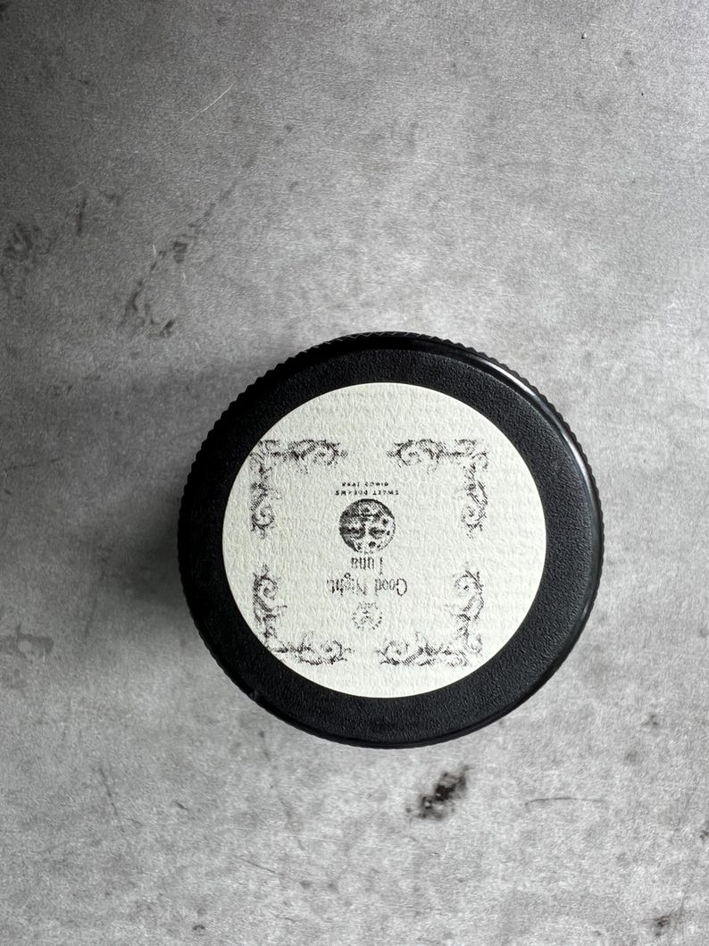 Op de afbeelding: Een ronde zwarte deksel van een potje met een cr&egrave;mekleurig etiket met sierlijke zwarte filigraanranden. Het etiket toont een centrale illustratie van een maan, de tekst "Good Night Luna" en de kleine tekst "SWEET DREAMS SINCE 1998".