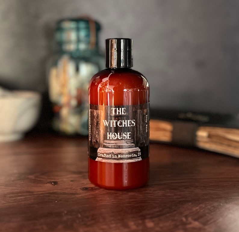 An Amber colored bottle of body wash labeled Witches House, crafted in Monrovia,In sits in from of a blurred background that includes a grimoire and mortal and pestle.