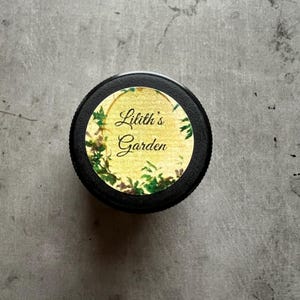Op de afbeelding: Een ronde zwarte deksel met een etiket op een gele achtergrond, versierd met groene klimopillustraties. De tekst Lilith's Garden is in een zwart cursief lettertype in het midden van het etiket gedrukt, omrand door een dunne gouden rand.
