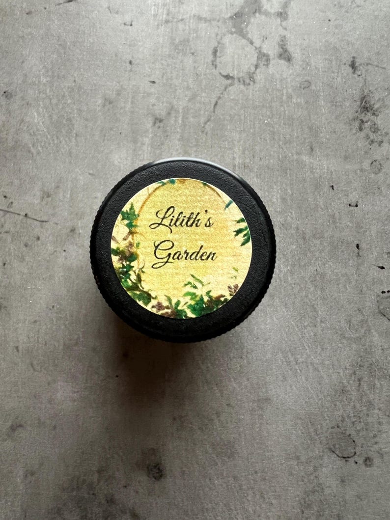 Op de afbeelding: Een ronde zwarte deksel met een etiket op een gele achtergrond, versierd met groene klimopillustraties. De tekst Lilith's Garden is in een zwart cursief lettertype in het midden van het etiket gedrukt, omrand door een dunne gouden rand.