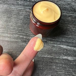 Op de afbeelding: Een pot gele cr&egrave;me met een wervelontwerp. Een kleine hoeveelheid van de cr&egrave;me zit op een vinger. De pot is bruin met een donkerbruin deksel. De achtergrond is een donkergrijze houtnerf.