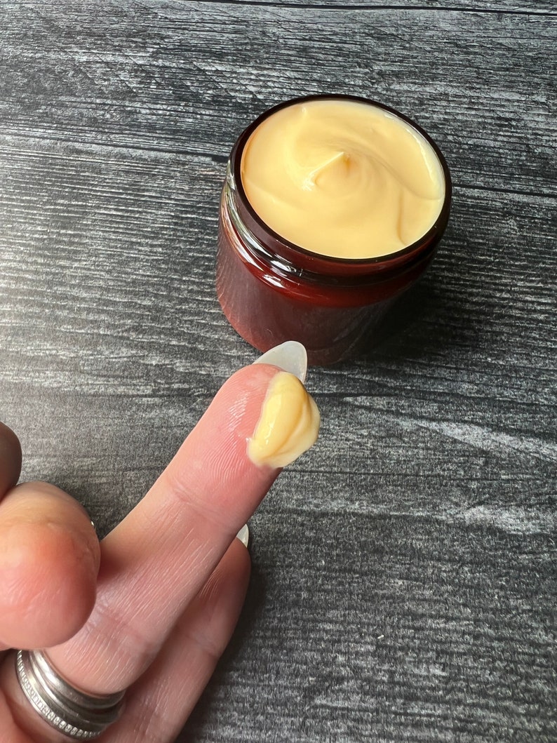 Op de afbeelding: Een pot gele cr&egrave;me met een wervelontwerp. Een kleine hoeveelheid van de cr&egrave;me zit op een vinger. De pot is bruin met een donkerbruin deksel. De achtergrond is een donkergrijze houtnerf.