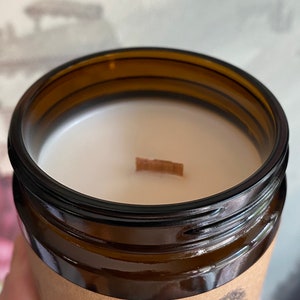 May include: Close-up of a brown glass candle jar filled with white wax. A wooden wick sits centered in the wax. The jar has a dark brown rim and a paper label around the base. The candle is likely scented.