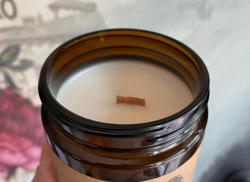May include: Close-up of a brown glass candle jar filled with white wax. A wooden wick sits centered in the wax. The jar has a dark brown rim and a paper label around the base. The candle is likely scented.