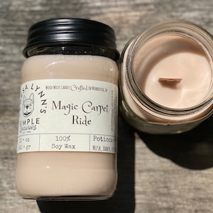 A hand holds a jar candle labeled Magic Carpet Ride with a Vintage Rug in the background. Label reads 100% Soy Wax Musk, Amber, Oud.  An open candle shows a sand colored wax with a wood wick.
