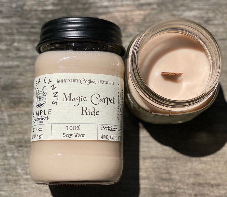 A hand holds a jar candle labeled Magic Carpet Ride with a Vintage Rug in the background. Label reads 100% Soy Wax Musk, Amber, Oud.  An open candle shows a sand colored wax with a wood wick.