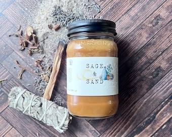 Pode incluir: Um frasco de vela Sage & Sand com tampa preta e rótulo branco. A vela é cercada por lavanda seca, um feixe de sálvia e um pedaço de palo santo. O frasco está em uma superfície de madeira.